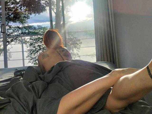 ScarlettN is lying on a bed with gray sheets, back to the camera, showing tattoos and a red headscarf, sunlit through a window, legs up, butt visible.