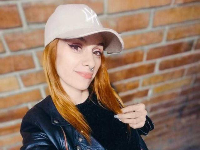 ScarlettN with red hair, wearing a beige NY cap, black leather jacket, and nose ring, smiling slightly, holding her hair. Brick wall in background.