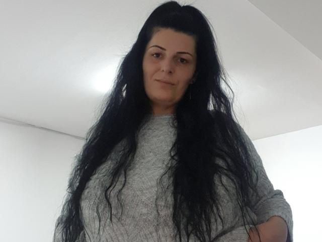 Mirim69 with long, wavy black hair, gray sweater, and a slight smile, looking down at the camera. Boobs are not visible. Her hands are in her pockets.