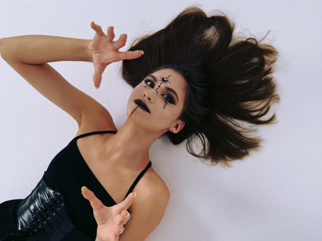 EllaCurtise with dark makeup, black lipstick, and star patterns on her face, lying down with arms raised, wearing a black top and black corset, brown hair splayed.