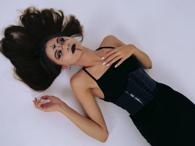 EllaCurtise laying on white, black makeup, dark lipstick, black dress, corset, black eye mask, long brown hair, hand on chest, corseted waist, pale skin, sexy.