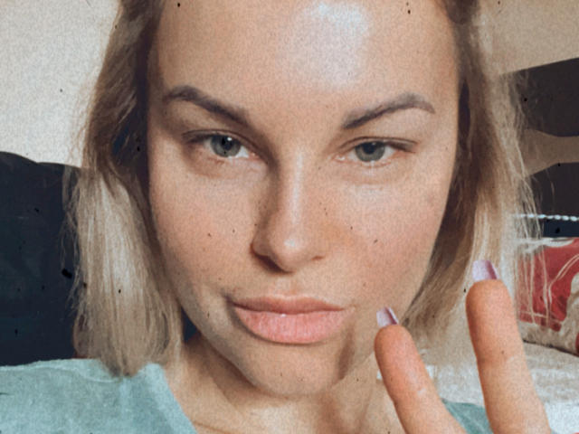 BelleLisaG has light skin, blonde hair, and green eyes with slightly pouty lips. She's making a peace sign with purple nails, wearing a green shirt.