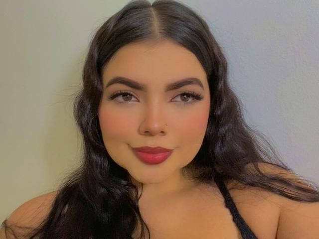 LaraJames has long black hair, brown eyes, red lipstick, and a small nose ring. She's wearing a black top, smiling with a cute face and a slightly curvy build.