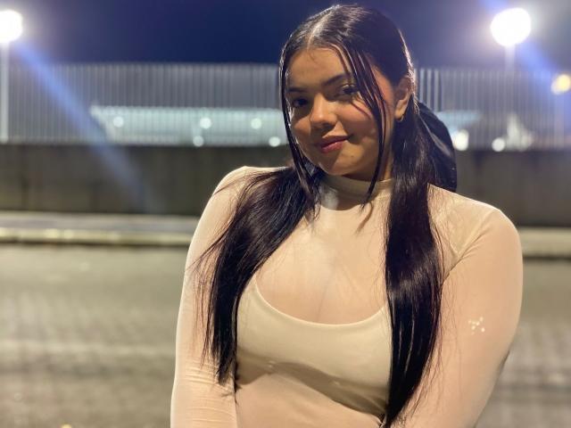 LaraJames with long black hair in pigtails, wearing a sheer white long-sleeve top, showing small tits, standing in a dark night setting with street lights.