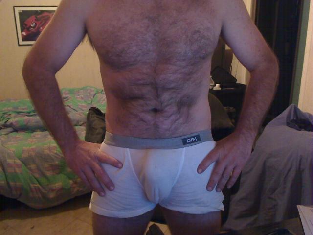 DomEric's hairy chest and abs, wearing white underwear with a visible bulge, hands on hips, in a dimly lit room. Bearded, shirtless, standing with back to camera.