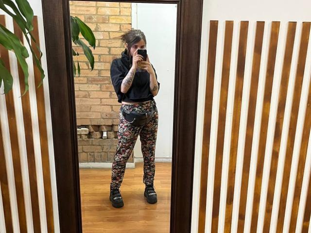 AmySpider standing in front of a mirror, wearing floral pants, black top, tattoos on arms, black shoes, holding phone, wooden floor, vertical wooden slats, plant on left.