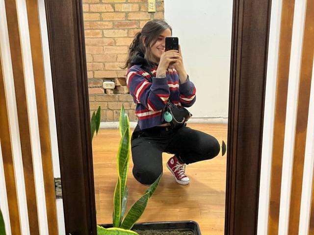 AmySpider, squatting, takes a mirror selfie, wearing a striped hoodie, black pants, red sneakers, holding a phone, with a green plant and wooden floor in the background.