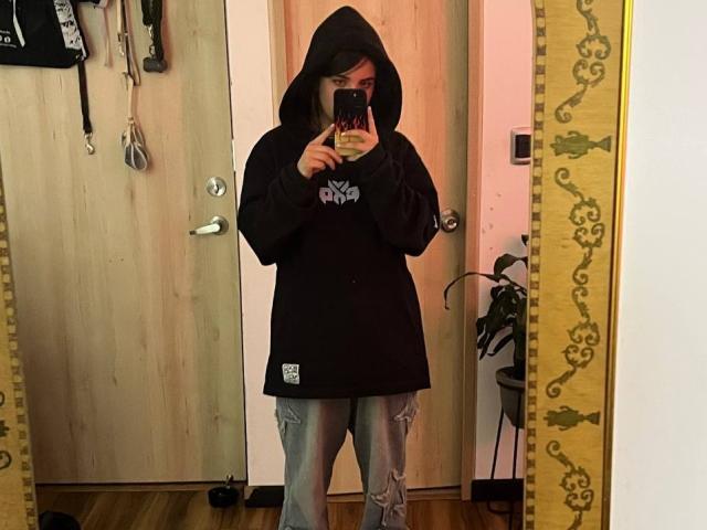 AmySpider, wearing a black hoodie with a white logo and ripped gray jeans, stands in front of a mirror, taking a selfie with a phone, covering her tits.