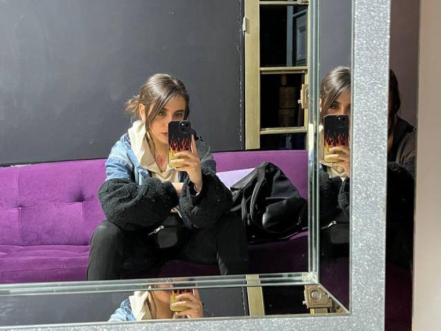 AmySpider takes a mirror selfie, showing her ass in black pants, with a blue jacket and black bag on a purple couch, holding a phone with a fiery design.
