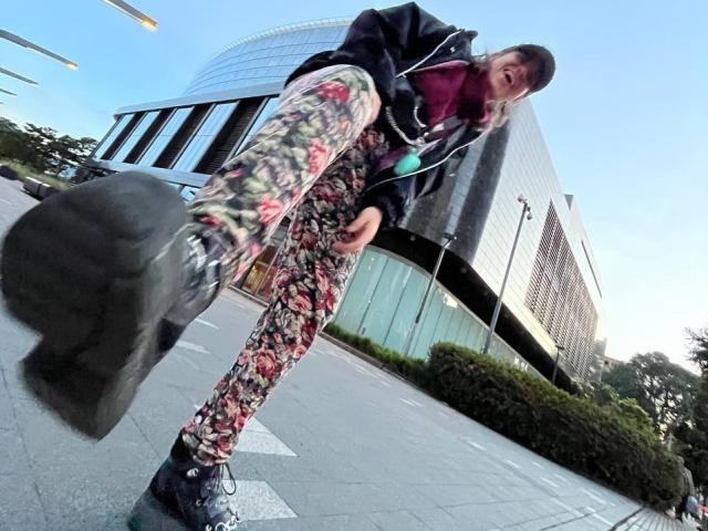 AmySpider with floral pants, black jacket, and purple mask, stepping on a black bag, low angle, blue sky, urban setting, black shoes, street, AmySpider's face slightly blurred.