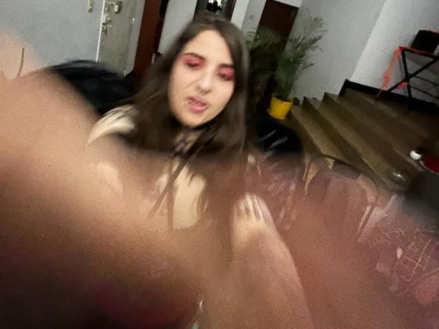 AmySpider with long brown hair, pink eyeshadow, and a nose ring, looking slightly off-camera, wearing a white top, with blurry foreground and dark background.