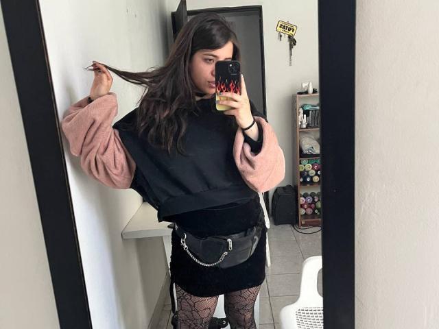 AmySpider with long dark hair, pink and black sweater, black mini skirt, fishnet stockings, and a black chain purse, taking a mirror selfie, holding hair.