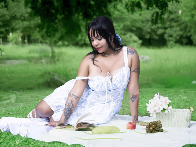 JessicaFiore with black hair, tattoos, and medium tits, wearing a white floral dress, lying on a blanket, reading a book, with grapes, apple, and flowers in a green field.