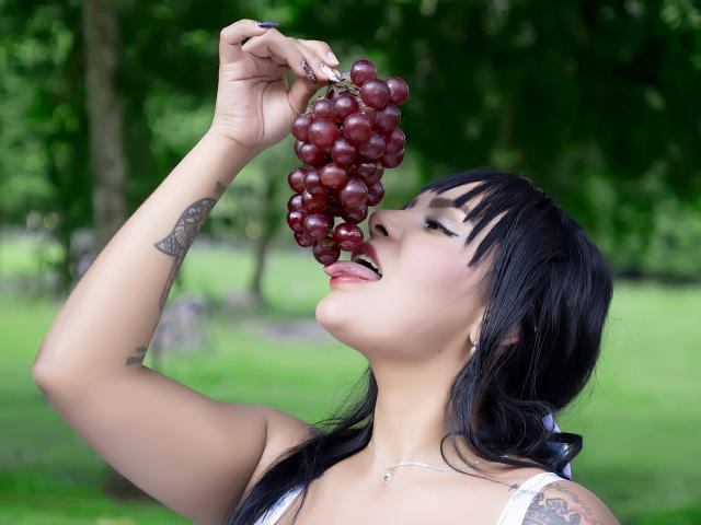 Jessica Fiore, con cabello oscuro y flequillo, sosteniendo un racimo de uvas rojas cerca de su lengua, con los ojos cerrados y la lengua afuera, tiene un tatuaje en su brazo derecho, en un parque verde..