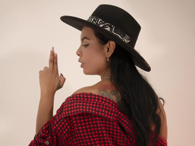 JessicaFiore with long black hair, red checkered shirt, off-shoulder, black hat with white pattern, tattoo on right shoulder, holding up middle finger, looking side profile.