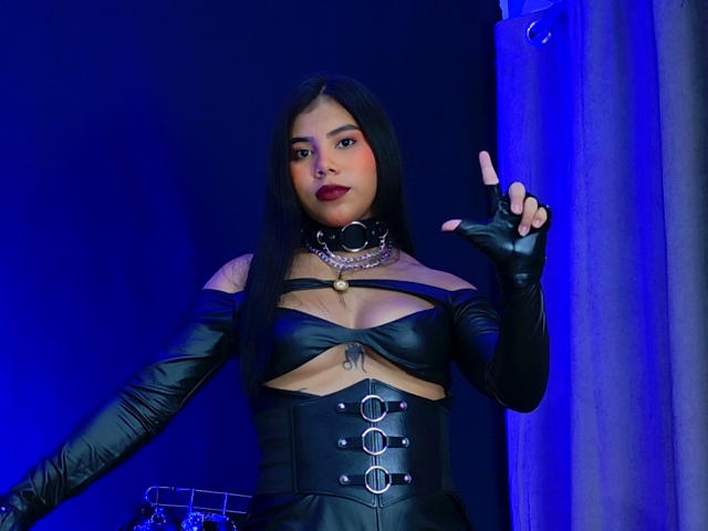 AmaniStone with long black hair, dark red lipstick, wearing a black leather outfit with cutouts, gloves, and a collar, raising her right finger, showing off her tits.