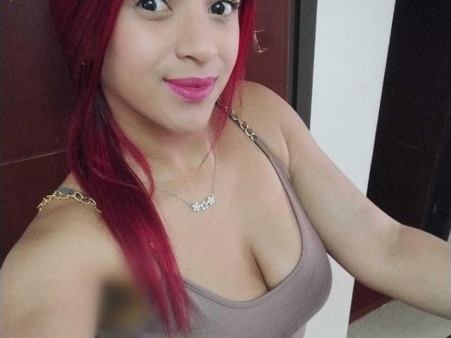 SweetieAlice69 has red hair, pink lipstick, and is wearing a low-cut gray tank top showing her big boobs. She's smiling and wearing a necklace with a small pendant.