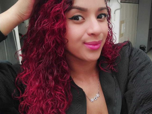 SweetieAlice69 with curly red hair, black zip-up jacket, pink lipstick, and winged eyeliner, wearing a silver necklace, smiling slightly, hand in hair.