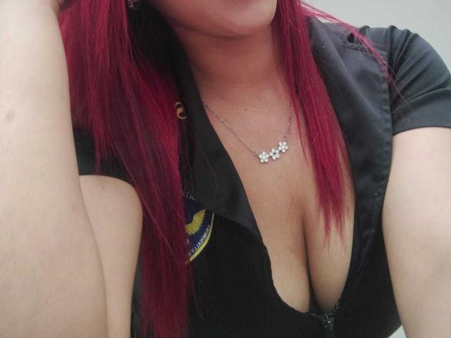 SweetieAlice69 with red hair, black shirt, deep cleavage, showing her big tits, wearing a flower necklace, her arm partially in frame, slightly bent.