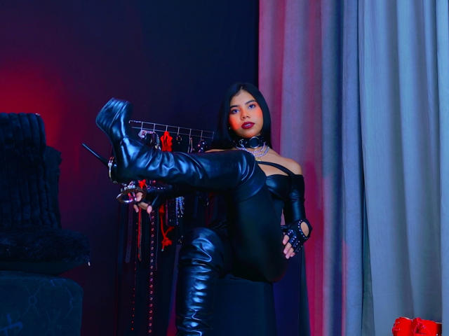 AmaniStone in black leather outfit, boots, and choker, lifting leg, showing black stiletto heel, with red lipstick, black hair, and serious expression, in dimly lit room.