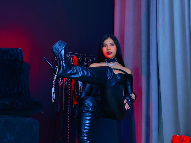 AmaniStone with black leather boots, off-shoulder top, and choker, raising one leg, showing her shiny boots, and red lips, standing next to BDSM gear in a dim room.