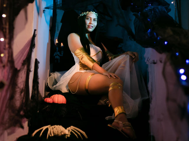 ValentinaMoreau with long black hair, gold arm bands, and white dress, sitting on black fabric, showing her tan thighs and gold leg straps, with a pink dildo in front.