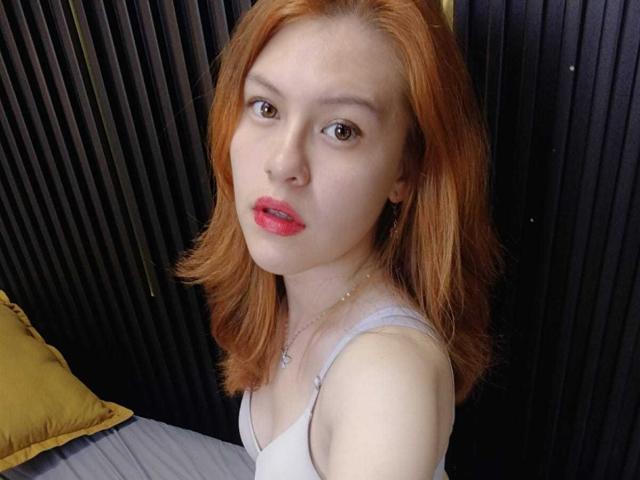 TaylorCarpert has pale skin, red hair, and red lips. She's wearing a white tank top, looking up with brown eyes, and has small, perky tits.