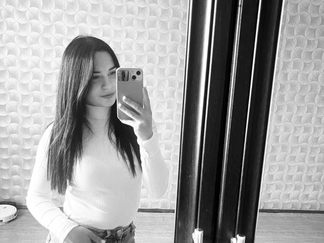 Black-and-white selfie of KelliValerie with long dark hair, white turtleneck, holding phone, standing in front of mirror, showing her small tits and subtle smile.