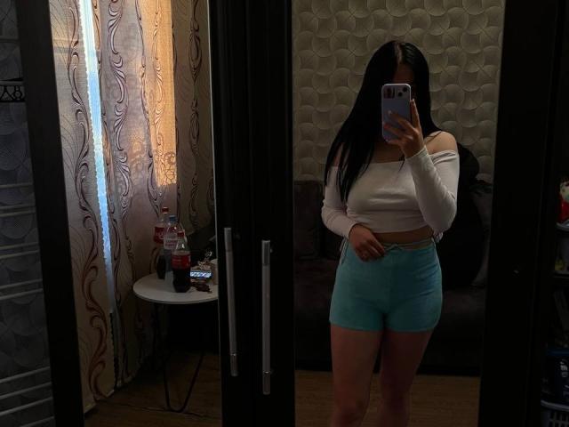 KelliValerie with long black hair, white off-shoulder top, and blue shorts, standing in front of a mirror, holding a phone, with a bottle and small table in background.