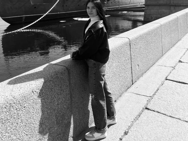 Black-and-white photo of KelliValerie standing by a concrete wall, wearing a dark jacket, jeans, and sneakers, with her hand on the wall, shadow on the ground.