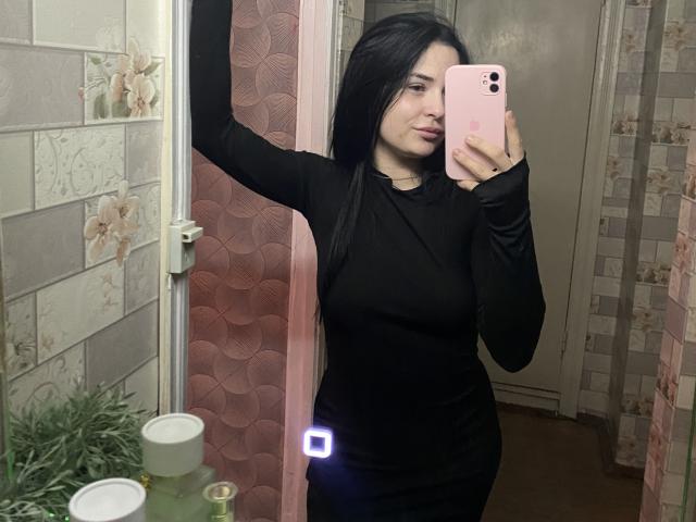KelliValerie with long black hair, wearing a tight black long-sleeve dress, taking a mirror selfie with a pink phone, showing her tits and a small butt light.