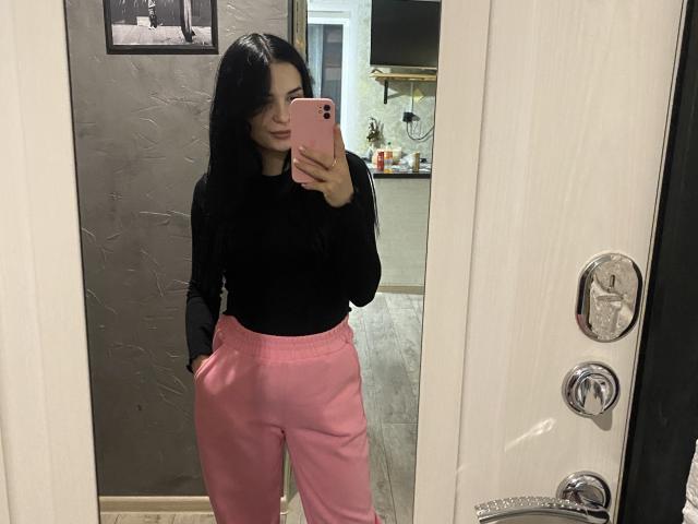 KelliValerie with long black hair, black long-sleeve shirt, pink pants, and pink phone, taking a mirror selfie, showing her ass, in a modern room.