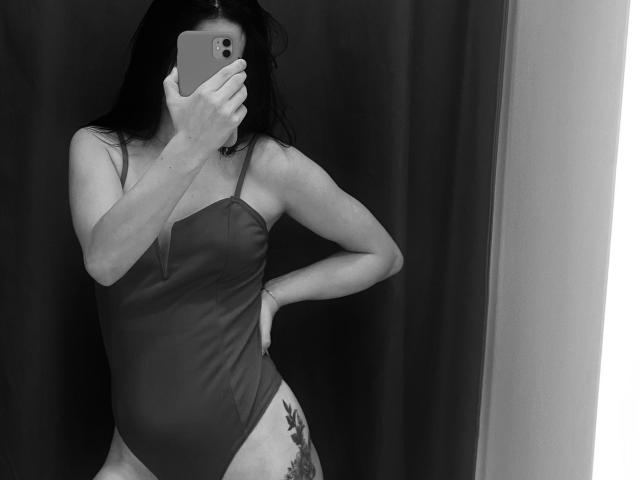 KelliValerie with wet black hair, wearing a strapless, dark one-piece, showing her ass and tattooed thigh, taking a mirror selfie with her phone, covering her face.