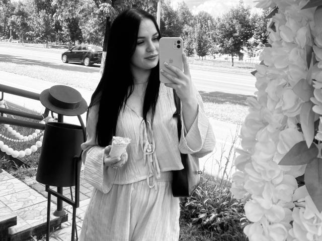 Black-and-white photo of KelliValerie with long dark hair, wearing a light dress and jacket, holding a cup, taking a selfie with an iPhone, standing near a flower-covered wall.