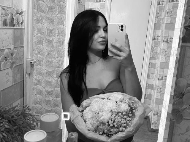 Black-and-white selfie of KelliValerie with long dark hair, holding a bouquet of flowers, standing in a bathroom, showing her bare boobs, holding an iPhone, mirror reflection.