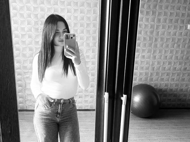 Black-and-white selfie of KelliValerie with long dark hair, white turtleneck, and high-waisted jeans, holding a phone, standing in front of a mirror, with a ball in the background.
