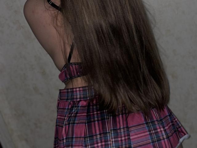 Madrinia's long brown hair covers her pink and black plaid dress, showing her bare back and black strap. Her ass is partially visible.