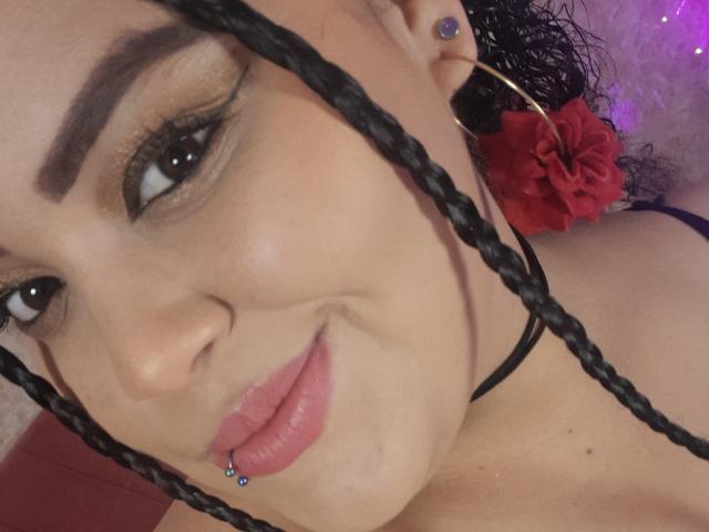 O rosto de Melannie Carter, com cabelo preto trançado, olhos escuros, sobrancelhas grossas, batom rosa e um piercing no nariz. Ela está usando um colar preto e uma flor vermelha no cabelo..