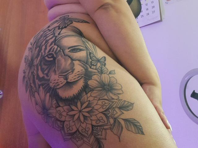 MelannieCarter's tattooed ass features a detailed black-and-gray tiger with flowers and butterflies, her hand resting on her hip, purple lighting in the background.