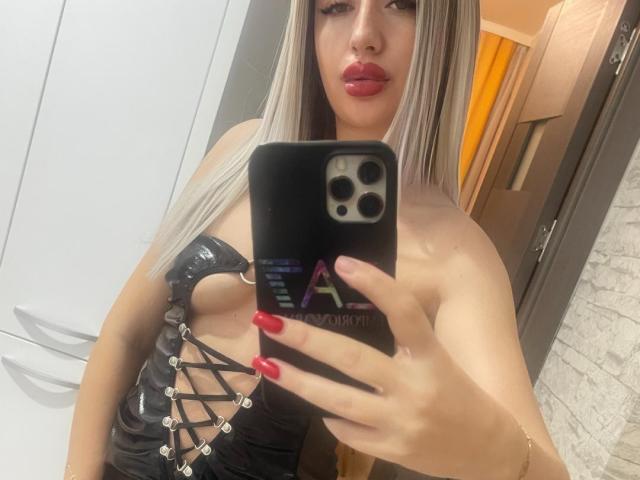 VanesaTop with long blonde hair, red lipstick, and red nails, taking a selfie in a mirror. She's wearing a black lace-up leather dress, exposing her tits.