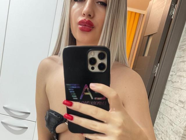 VanesaTop with red lipstick and long blonde hair, holding a black phone with red nails, taking a mirror selfie, showing her bare shoulders and black scrunchie.