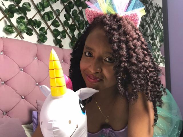 ArtemisaS with curly brown hair, wearing colorful cat ears and a purple top, smiles at a yellow-striped unicorn plushie, sitting on a pink tufted couch.