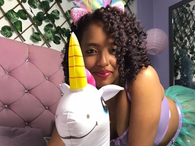 ArtemisaS with curly brown hair, purple lingerie, and a unicorn headband, leaning on a yellow unicorn pillow, smiling at the camera. She has medium brown skin and pink lipstick.