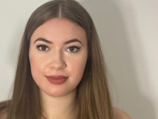 LeaHoty has long brown hair, brown eyes, and is smiling with red lipstick, showing her soft, fair skin and full lips. She's looking directly at the camera.