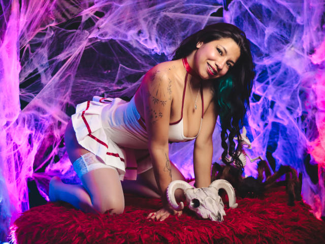 PaulinaColemann on all fours, wearing a white nurse outfit with red trim, white stockings, and a red collar, smiling at the camera, with a ram skull and red fur on the floor.