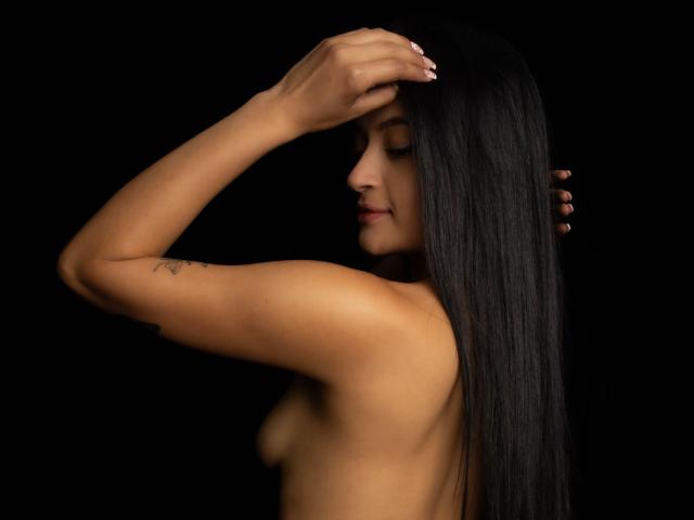 LizzyMarhall69 with long black hair, topless, arm raised, hand on head, small tattoo on arm, dark background, subtle smile, small tits, smooth skin, dark brown eyes.