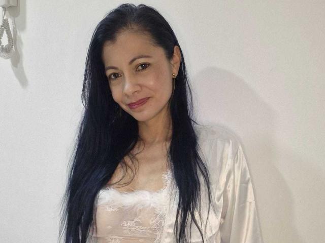 MelissaBaud with long black hair, wearing a white lace top and silky robe, smiling at the camera. Her tits are small, she's got cute brown eyes, and red lipstick.