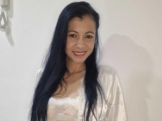 Melissa Baud, avec ses longs cheveux bleu foncé, souriante, portant un haut en dentelle blanche et une robe en soie, laissant entrevoir de petites seins. Lumière douce, fond blanc, petit appareil fixé au mur sur la gauche..