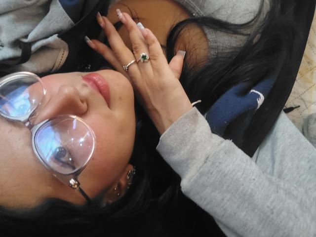 VioletXBloom with glasses, dark hair, and pale skin, lying down, hand near face with green ring, gray sweater, and pink lips, looking at camera. Boobs partially visible.