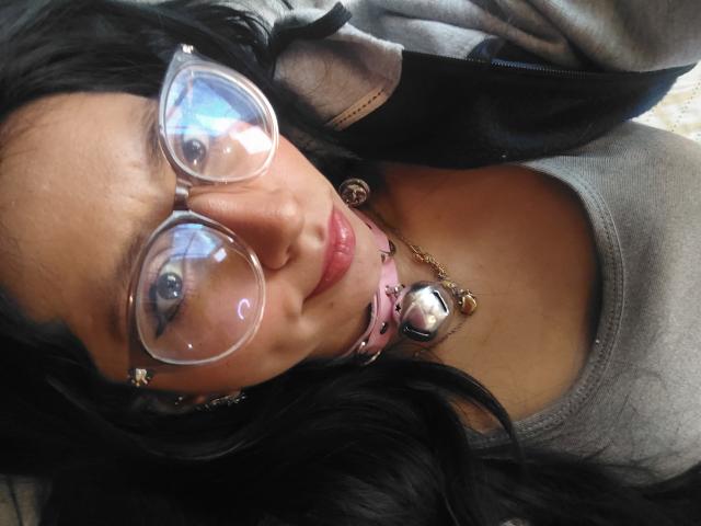 VioletXBloom has dark hair, glasses, pink lips, and a cute face with a necklace. She's wearing a gray top and a black jacket. Her tits are small and cute.